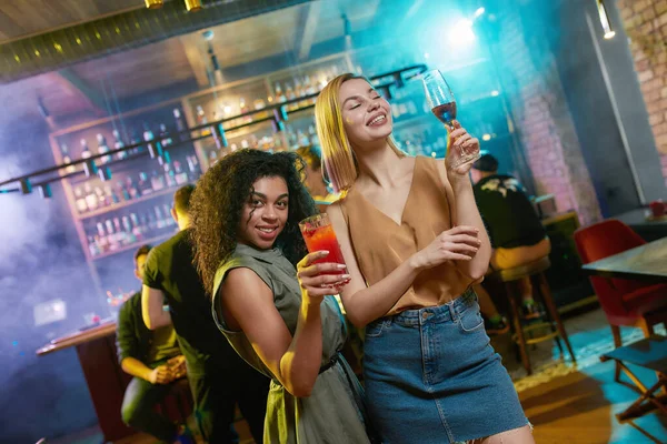 Mujeres jóvenes atractivas emborrachándose, posando con un cóctel en sus manos. Amigos celebrando, divirtiéndose en el bar — Foto de Stock