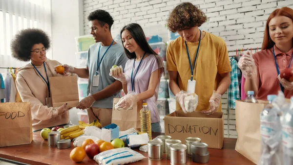 Young male and female volunteers packing food and drinks donation for homeless into boxes and paper bags, Small group of people working in charitable foundation