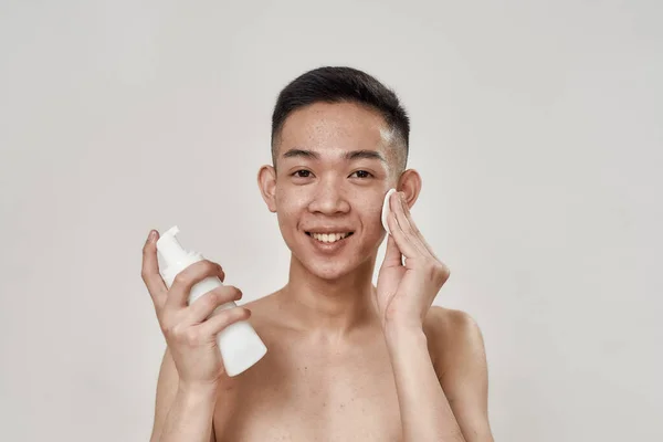 Porträtt av tröjlös ung asiatisk man med problematisk hud rengöring ansiktet med lotion med hjälp av bomullspads, ler mot kameran isolerad över vit bakgrund — Stockfoto