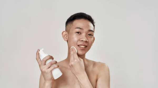 Portrait of shirtless young asian man with problematic skin cleaning his face with lotion using cotton pads, looking at camera isolated over white background