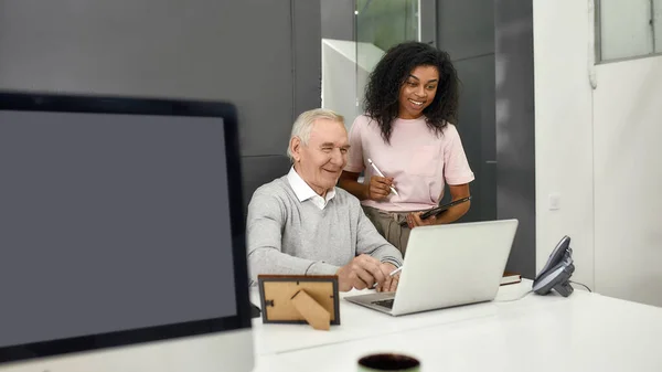 Aged man, senior intern looking at laptop while showing results to his young colleague, Friendly female worker mentoring and training new employee, monitoring his progress at work — Stock Photo, Image