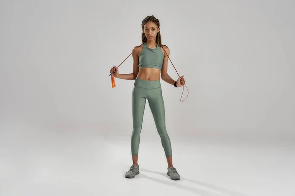 Ganzkörperaufnahme einer jungen athletischen, schlanken Mixed Race Frau in Sportbekleidung, die vor grauem Hintergrund steht und Springseil hält — Stockfoto