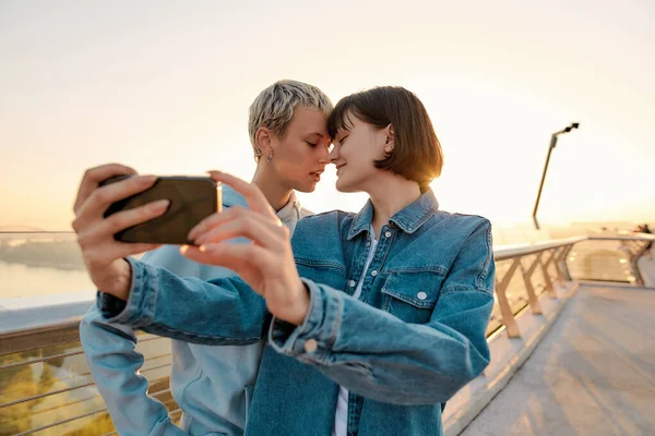 Couple lesbien debout sur le pont, posant tout en prenant une photo selfie, regardant le lever du soleil ensemble — Photo