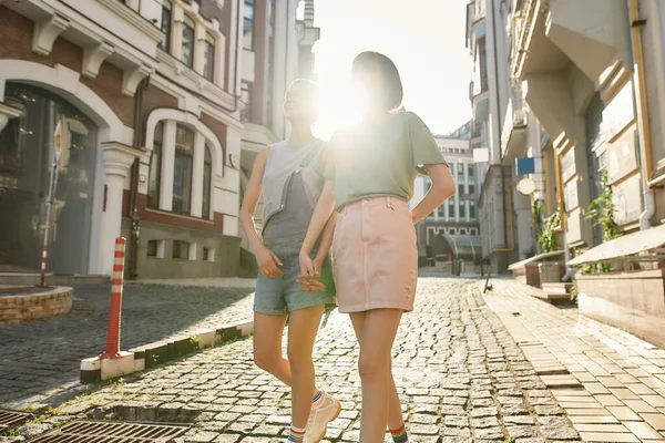 Pareja joven lesbiana, dos mujeres en ropa casual cogidas de la mano, caminando por la calle mientras exploran la ciudad en un día soleado — Foto de Stock