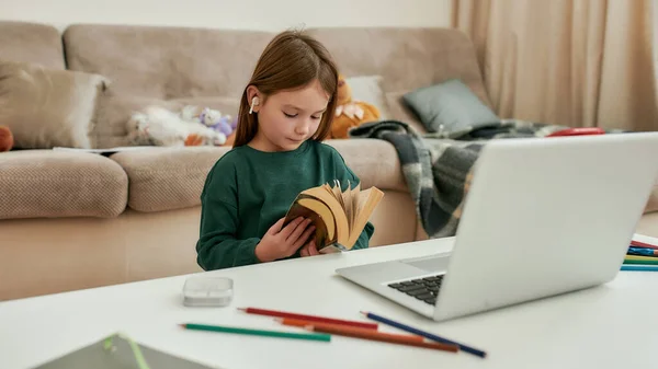 Uma menina pequena rolando um livro enquanto se senta sozinha na frente de um laptop em uma mesa durante a educação à distância — Fotografia de Stock