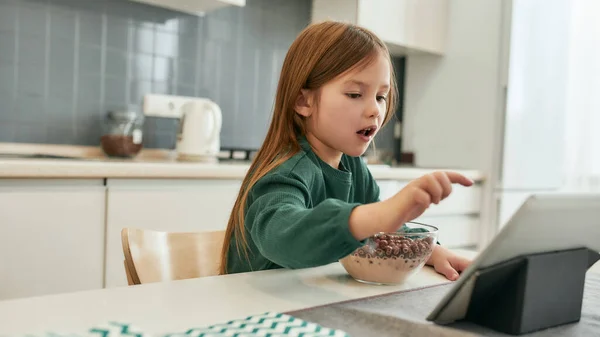 Uma menina está batendo em uma tela de tablet enquanto come suas bolas de cereais rápidas com leite sentado à mesa em uma grande cozinha brilhante — Fotografia de Stock