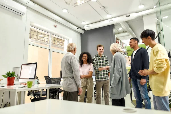 Vidám fiatal csapat köszönti az új alkalmazottakat, idős férfit és nőt, idősebb gyakornokok kezet ráznak a kollégákkal a modern irodában — Stock Fotó
