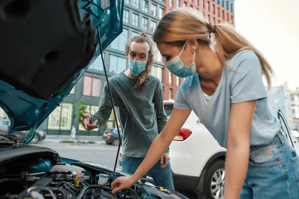 Hombre con máscara médica ayudando a chica con su auto mientras ella está examinando, mirando su coche roto con capucha abierta, de pie en la calle de la ciudad — Foto de Stock