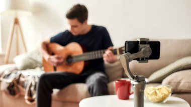 Genç adam evde gitar çalarken akıllı telefon kullanıyor ve video kaydediyor. Blogculuk, teknoloji konsepti. Bir tripoda cep telefonuna odaklan