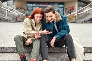 İki genç ellerinde akıllı telefonlar, ekrana bakıyor, bir şeyleri tartışıyor ve merdivenlerde otururken gülümsüyor.