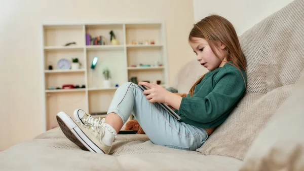 Una linda niña pequeña sosteniendo la tableta sentada en un sofá frente a las estanterías —  Fotos de Stock