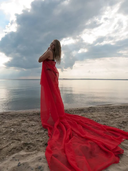 A young female dancer with a slender body in a black bikini and a red veil on the beach against a stormy sky