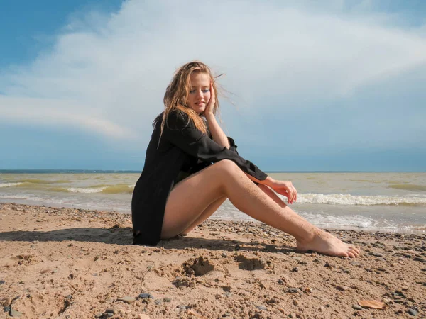 A young beautiful woman with loose blonde hair in a black shirt and a bikini with full thighs on the sandy beach against the background of the sea surf on a Sunny day