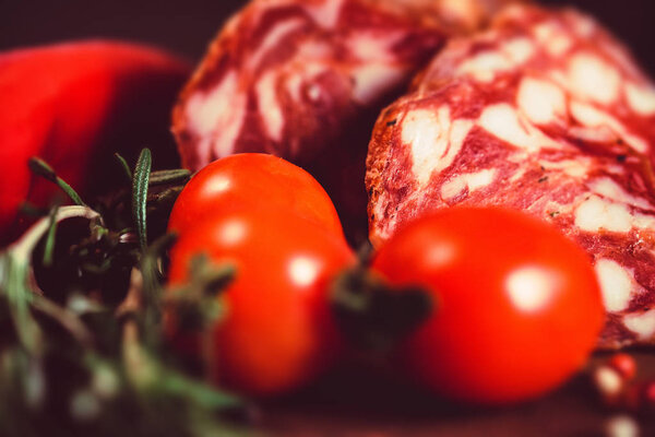 Close up image of cured sausage on wooden board with fresh tomatoes, herbs and peppercorn. Selective focus