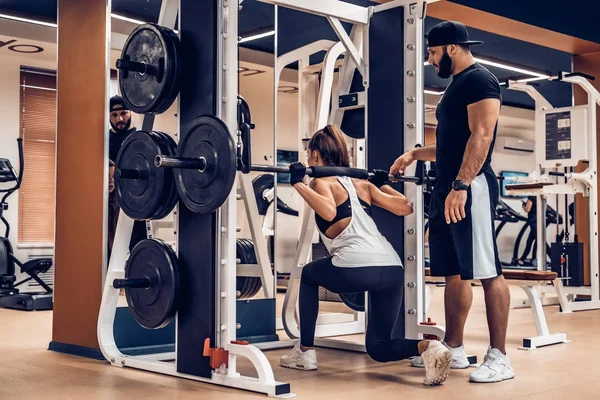 Atractiva joven mujer hace sentadillas de barra con entrenador en el gimnasio — Foto de Stock