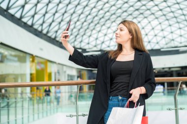Alışveriş poşetleri ile güzel kahverengi saçlı kız alışveriş merkezinde selfie alır