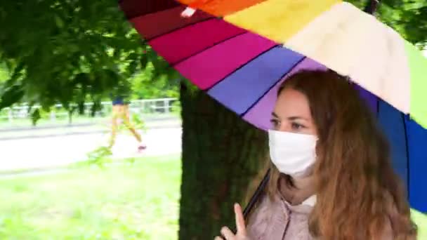 Chica caucásica en una máscara protectora camina bajo un paraguas en una calle vacía bajo la lluvia de primavera. Seguridad y distancia social durante una pandemia de coronavirus. Nueva normalidad, las implicaciones de la cuarentena. — Vídeo de stock