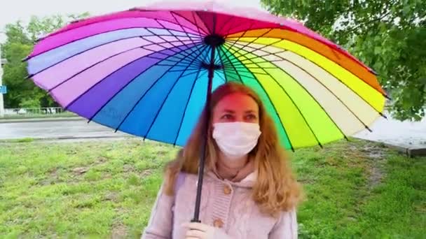 Caucasian girl in a protective mask walks under an umbrella on an empty street in spring rain. Safety and social distance during a coronavirus pandemic. New normal, the implications of quarantine. — Stock Video