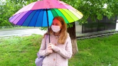 Koruyucu maskeli beyaz kız bahar yağmurunda boş bir sokakta şemsiye altında yürüyor. Koronavirüs salgını sırasında güvenlik ve sosyal mesafe. Yeni normal, karantinanın sonuçları..