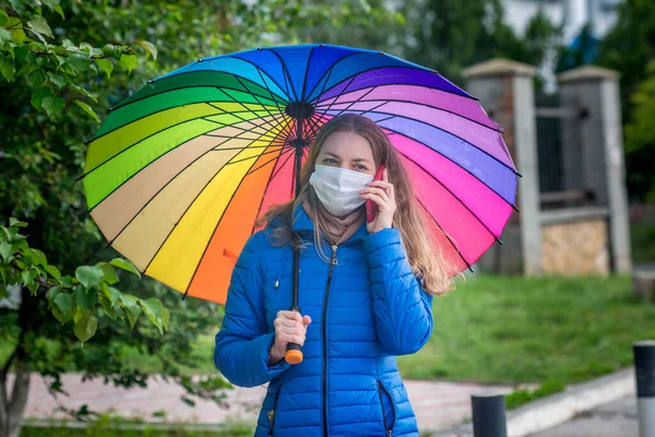 Uma menina caucasiana com uma máscara protetora fica em uma rua vazia em uma parada de ônibus sob um guarda-chuva na chuva de primavera e fala ao telefone. Segurança e distância social durante uma pandemia de coronavírus . — Fotografia de Stock