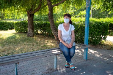 Koruyucu maskeli yetişkin bir kadın boş bir otobüs durağında tek başına oturur ve toplu taşıma araçlarını bekler. Sosyal mesafe. Otobüs yolcuları koronavirüsten korunuyor. Yeni kural. Bir salgın sırasında yaşam biçimi