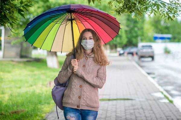 A menina caucasiana em uma máscara protetora anda abaixo de um guarda-chuva em uma rua vazia na chuva de primavera. Segurança e distância social durante uma pandemia de coronavírus. Novo normal, as implicações da quarentena. — Fotografia de Stock