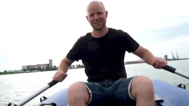 Un joven guapo está flotando en el río en un bote de goma. El pescador está remando en el mar y sonriendo alegremente. Actividades al aire libre, excursiones en barco de verano y pesca . — Vídeo de stock