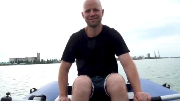 Un jeune bel homme flotte sur la rivière dans un bateau en caoutchouc. Le pêcheur rame dans la mer et sourit joyeusement. Activités de plein air, excursions en bateau d'été et pêche. — Video