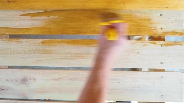 Ζωγραφίζοντας έναν ξύλινο πάγκο με προστατευτικό ανθεκτικό στις καιρικές συνθήκες χρώμα. Το γυναικείο χέρι εφαρμόζει ένα κίτρινο εμποτισμό με ένα πινέλο. Επεξεργασία επιφάνειας από έντομα. Η έννοια του DIY. Κλείσε.. — Αρχείο Βίντεο