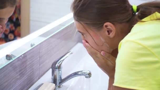 Una joven se lava la cara con agua limpia en el baño. El adolescente se ríe alegremente y se lava el jabón. Procedimientos de higiene matutina y nocturna. — Vídeo de stock