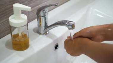Genç bir çocuk lavaboda akan suyun altında ellerini sabunla yıkıyor ve sonra musluğu kapatıyor. Bakteri ve virüs bulaşmasını önlemek için ellerinizi temizleyin. Bir salgın sırasında koruma.