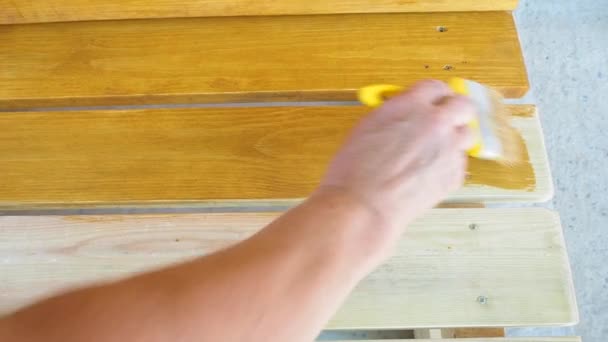 Pintar un banco de madera con pintura protectora resistente a la intemperie. La mano femenina aplica una impregnación amarilla con un pincel. Tratamiento superficial de insectos. El concepto de bricolaje. De cerca.. — Vídeo de stock