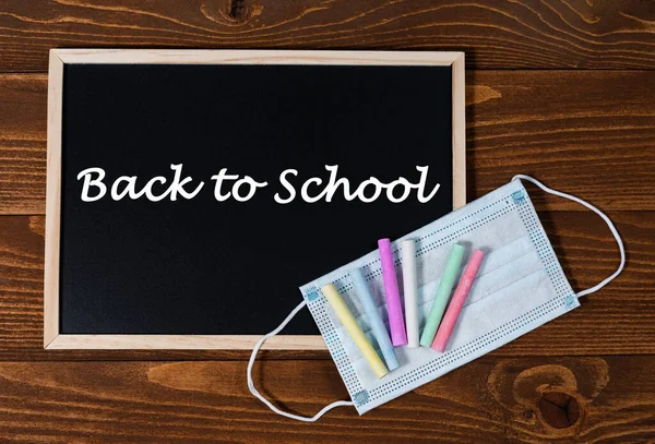 A blackboard with text Back to School, set of colored crayons and a protective mask on a wooden background. Start of school year and sale of school supplies. Home education and learnin after pandemic.