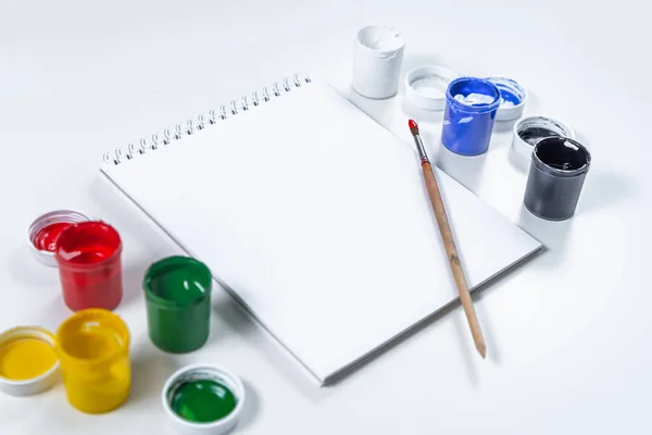 Artistic mock-up on a white background with space for text. Drawing tools: colored acrylic paint, paintbrush, and a blank Notepad layout. Artists table with accessories. Home education and training.
