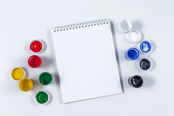 Artistic mock-up on a white background with space for text. Drawing tools: colored acrylic paint, paintbrush, and a blank Notepad layout. Artists table with accessories. Home education and training. — Stock Photo, Image