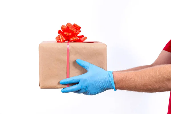 A koronavírus-világjárvány idején az ajándékok biztonságos, érintésmentes, távoli szállítása. Közelről. Futár kéz védő orvosi kesztyű tart egy szép ajándék doboz egy íj egy fehér háttér. — Stock Fotó