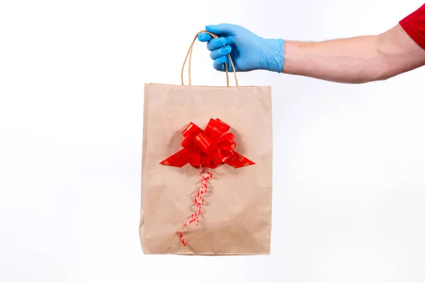 在大肠病毒大流行期间，确保非接触式远程递送节日礼物。靠近点送礼人手戴着防护医用手套，手里拿着一个白色背景上有蝴蝶结的纸袋. — 图库照片