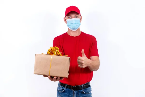 Bezpečná dodávka dárků na dovolenou. Kurýr v červené uniformě a ochranné lékařské masce drží krabici s lukem. Bezkontaktní dálkové dárkové objednávky v karanténě během pandemie koronaviru. — Stock fotografie