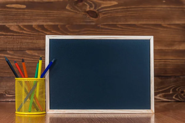 Um quadro negro com espaço para texto, um conjunto de lápis coloridos com um copo sobre um fundo de madeira. Início do conceito do ano letivo e venda de material escolar. Educação e aprendizagem em casa. — Fotografia de Stock