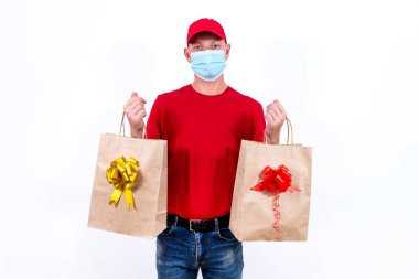 Coronavirus salgını sırasında uzak mesafeden tatil hediyelerini güvenli bir şekilde teslim etmek. Kırmızı üniformalı ve koruyucu tıbbi maskeli bir kurye beyaz arka planda fiyonklu iki güzel hediye paketi taşır..