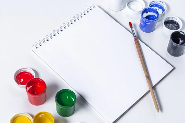 Artistic mock-up on a white background with space for text. Drawing tools: colored acrylic paint, paintbrush, and a blank Notepad layout. Artists table with accessories. Home education and training. — Stock Photo, Image