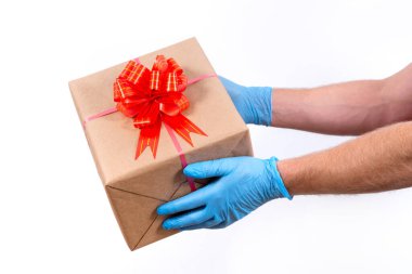 Coronavirüs salgını sırasında uzak mesafeden tatil hediyelerini güvenli bir şekilde teslim etmek. Kapatın. Koruyucu tıp eldivenleri içindeki kurye elinde beyaz arka planda fiyonklu güzel bir hediye kutusu var..