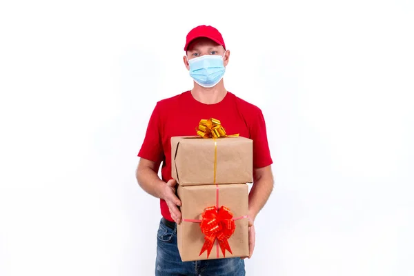 Безпечна дистанційна доставка святкових подарунків під час пандемії коронавірусу. Кур'єр в червоній формі і захисна медична маска тримає дві красиві подарункові коробки з бантами . — стокове фото