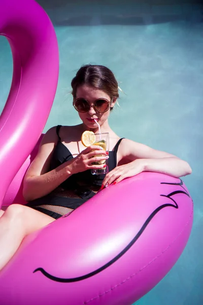 Genç ve güzel seksi kadın havuzda yüzüyor ve şişme pembe flamingoda kokteyl içiyor.