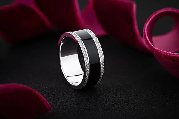 Female wedding silver ring with black enamel and diamonds on black background with red ribbon. Unique white gold ring band. Advertising jewelry still life