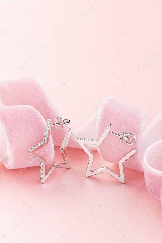 Star shaped silver earring studs decorated with diamonds on pastel pink background with ribbon. Jewelry still life 