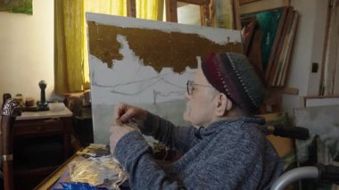 Tekerlekli sandalyedeki kıdemli sanatçı tuvaldeki yağlı boya resme gümüşü uygular.