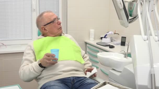 Старик сидит в стоматологической клинике и смотрит на часы, пьющие воду — стоковое видео