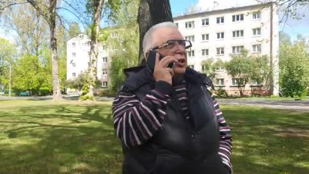 Gråhårig man talar på smartphone promenader i stadsparken — Stockvideo