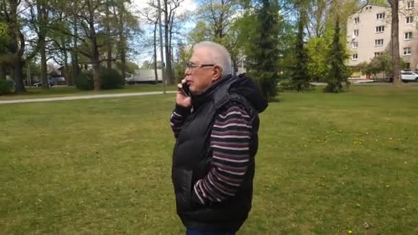 Senior man loopt langs groene gazon praten op mobiele telefoon — Stockvideo